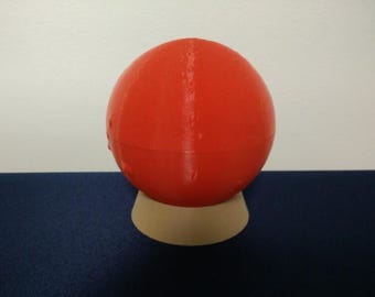 May include: A vibrant red sphere, possibly a container or ornament, rests on a beige, rounded base. The object's surface shows a slight texture and a vertical seam. The background is a simple white and navy blue.