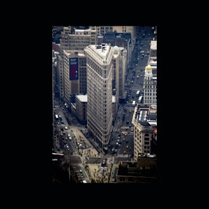 May include: An aerial view of a city street with a tall, narrow building in the centre. The building has a unique, stepped facade and is surrounded by other buildings. The street is lined with cars and pedestrians.