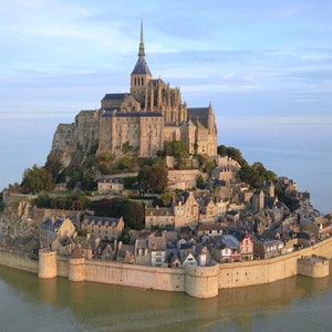 Może przedstawiać: Widok z lotu ptaka na Mont Saint-Michel, historyczne opactwo położone na wyspie pływowej w Normandii, we Francji. Opactwo wznosi się na skalistym cyplu i jest otoczone wodą podczas przypływu.
