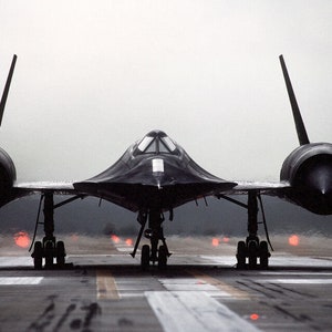 Puede incluir: Un avión negro SR-71 Blackbird está posicionado en una pista. El avión de reconocimiento de gran altitud tiene un diseño elegante y aerodinámico con prominentes góndolas de motor y estabilizadores verticales. El tren de aterrizaje es visible y las luces de la pista están encendidas.