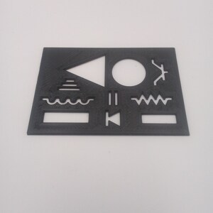 Op de afbeelding: Een zwarte plastic sjabloon met verschillende vormen en symbolen, waaronder een driehoek, een cirkel, golvende lijnen en een rechthoek.