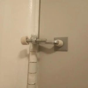 Puede incluir: Un tope de puerta blanco con un brazo de metal y un tope de goma blanco en cada extremo. El tope de puerta está montado en una pared blanca.