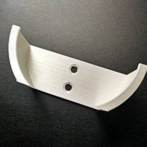 Pode incluir: Suporte de parede de plástico branco com dois furos para parafusos. O suporte tem uma forma curva em ambas as extremidades.