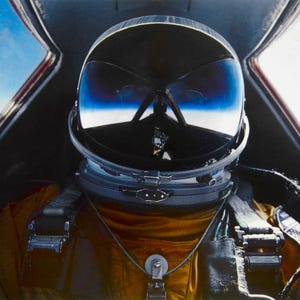 May include: A pilot in a black helmet with a reflective visor sits in the cockpit of a jet. The pilot wears a gold flight suit and is secured by straps. The cockpit frame is black, and the sky is visible through the canopy.