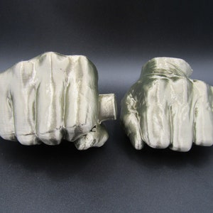 May include: Two silver-colored 3D printed fists, each with a closed fist shape. The fists are positioned side by side on a black surface.