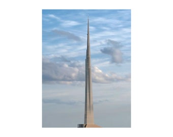 May include: A tall, slender, silver building with a pointed top against a blue sky with white clouds.