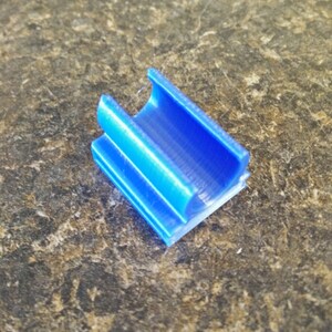 K&ouml;nnte beinhalten: Ein kleines, blaues Plastikteil mit einem Kanal, der durchl&auml;uft. Das Teil ist so konzipiert, dass es als Verbinder oder F&uuml;hrung verwendet werden kann.