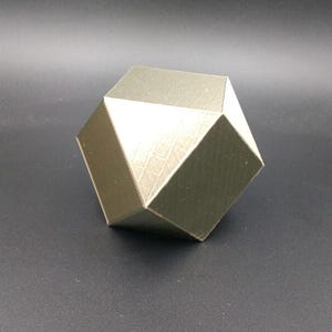 Op de afbeelding: Een metallic gouden geometrische sculptuur met een veelzijdig ontwerp. Het object heeft een getextureerd oppervlak en staat tegen een donkere achtergrond. De sculptuur heeft de vorm van een complex veelvlak.