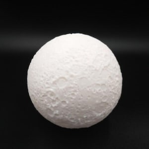 Könnte beinhalten: Ein weißes, kugelförmiges Dekorationsobjekt, das dem Mond ähnelt. Die Oberfläche ist mit Kratern und anderen Mondmerkmalen strukturiert. Das Objekt befindet sich vor einem schwarzen Hintergrund.