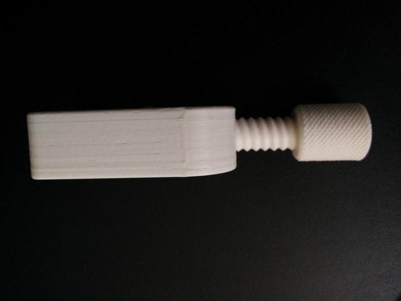 May include: A white plastic clamp with a threaded screw and a knob. The clamp is designed to hold objects together.