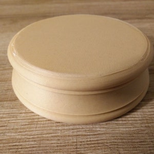 May include: A round, cream-colored wooden pedestal with a smooth, slightly textured surface. The pedestal has a raised lip around the top and bottom.