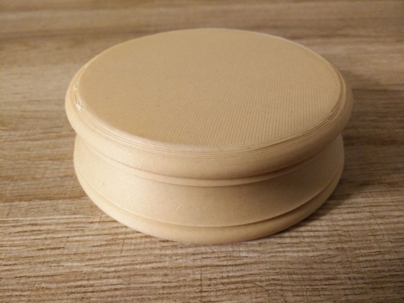 May include: A round, cream-colored wooden pedestal with a smooth, slightly textured surface. The pedestal has a raised lip around the top and bottom.