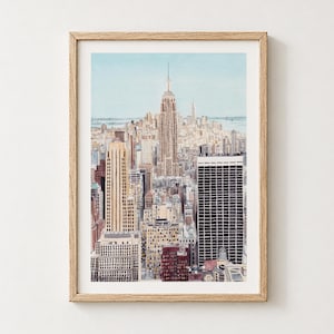 May include: A watercolor painting of the New York City skyline featuring the Empire State Building in the center, surrounded by various skyscrapers in shades of beige, brown, and black, all framed in a light-colored wooden frame.