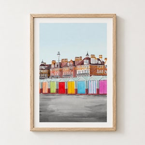 May include: A framed watercolor print depicting a row of colorful beach huts in front of multi-story brick buildings under a pale blue sky. The huts feature vertical stripes in orange, green, red, yellow, blue, and pink, set against a gray ground.