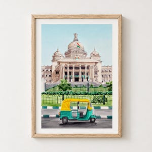 Puede incluir: Una pintura de acuarela en un marco de madera que muestra un mototaxi de color verde azulado y amarillo estacionado frente al edificio Vidhana Soudha, que presenta una gran cúpula central y una bandera india ondeando en la cima.