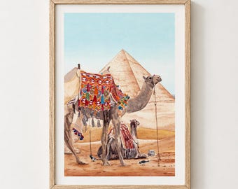 May include: A framed watercolor print depicting two camels in a desert landscape with a large pyramid in the background. One camel stands adorned with a colorful, patterned saddle blanket featuring red, blue, and yellow tassels, while another rests.