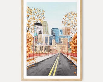 May include: A watercolor print in a light wood frame depicting a city skyline viewed from a road with yellow lane markings. The scene features autumn trees with orange and yellow leaves lining the street, leading toward tall buildings under a pale blue sky.