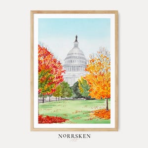 May include: A framed watercolor print depicting the United States Capitol building in Washington D.C. surrounded by autumn trees with vibrant red, orange, and yellow foliage. The scene features a green lawn with fallen leaves and a light blue sky.