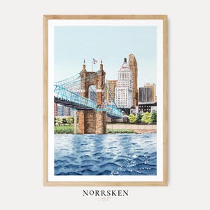 May include: Watercolor painting of a cityscape featuring a blue suspension bridge over a river, with buildings in the background. The artwork is framed in a light wood frame. The water is a deep blue, and the sky is a light blue. The artist's name, NORRSKEN Art, is at the bottom.