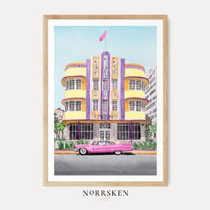 May include: A watercolor art print in a light wood frame depicting a yellow and purple Art Deco building with the word MARLIN on the facade. A long pink vintage car is parked in front of the building on a gray street under a clear blue sky.