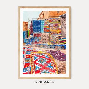 Op de afbeelding: Een aquarel in een lichthouten lijst met een levendige buitenmarktscène met talloze kleurrijke patronen tapijten en textiel die op rekken worden getoond en over de grond zijn verspreid. De tekst NORRSKEN Art staat eronder afgedrukt.