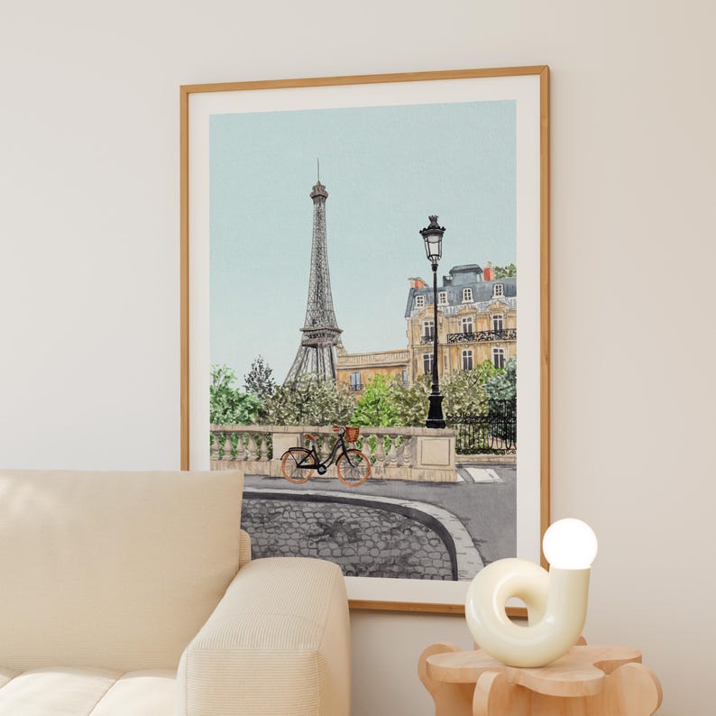 May include: Watercolor print of the Eiffel Tower in Paris, France, with a bicycle parked on a cobblestone street. The artwork features a light blue sky and a classic Parisian street scene, framed in a wooden frame.