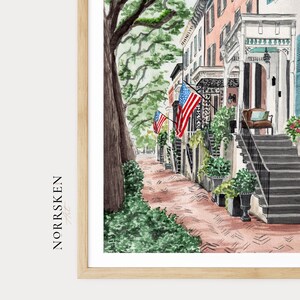 May include: A watercolor print in a light wood frame depicting a street scene with a large tree, a brick sidewalk, and historic buildings featuring porches and stairs. American flags are displayed on the buildings. The text NORRSKEN Art is visible.
