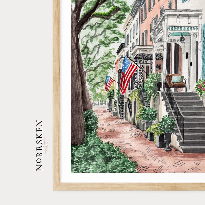 May include: A watercolor print in a light wood frame depicting a street scene with a large tree, a brick sidewalk, and historic buildings featuring porches and stairs. American flags are displayed on the buildings. The text NORRSKEN Art is visible.