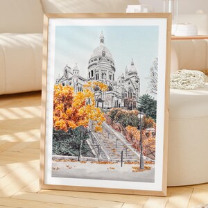 K&ouml;nnte beinhalten: Gerahmter Aquarelldruck der Basilika Sacr&eacute;-C&oelig;ur in Paris. Das Kunstwerk zeigt die Basilika mit blauem Himmel, Herbstb&auml;umen mit orangefarbenen Bl&auml;ttern und einer Treppe, die zur Kirche f&uuml;hrt. Der Rahmen ist aus hellem Holz.