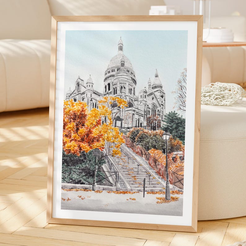 K&ouml;nnte beinhalten: Gerahmter Aquarelldruck der Basilika Sacr&eacute;-C&oelig;ur in Paris. Das Kunstwerk zeigt die Basilika mit blauem Himmel, Herbstb&auml;umen mit orangefarbenen Bl&auml;ttern und einer Treppe, die zur Kirche f&uuml;hrt. Der Rahmen ist aus hellem Holz.