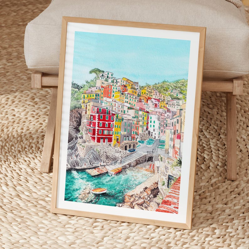May include: A framed watercolour painting depicting a coastal village with colourful buildings stacked on a cliffside overlooking a turquoise harbour with small boats. The artwork features vibrant red, yellow, and white structures, rocky terrain, and a clear blue sky.