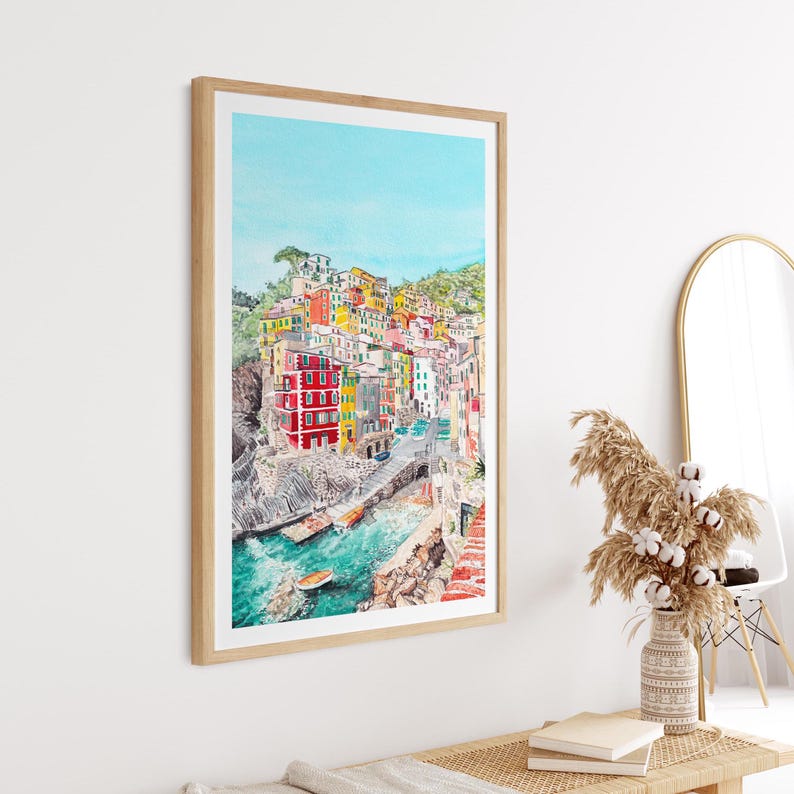 May include: A framed watercolour painting depicting a coastal village with colourful buildings stacked on a cliffside overlooking a turquoise harbour with small boats, all enclosed in a light-toned wooden frame hanging on a plain white wall.