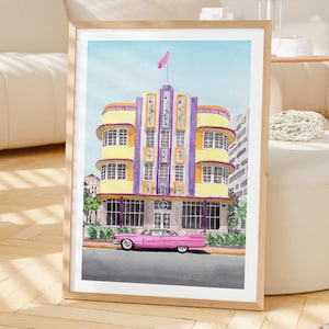 May include: A framed watercolor print depicting the Marlin Hotel in Miami with its yellow and purple art deco facade, featuring a pink vintage car parked in front. The building has multiple windows, a central tower, and a pink flag on top.