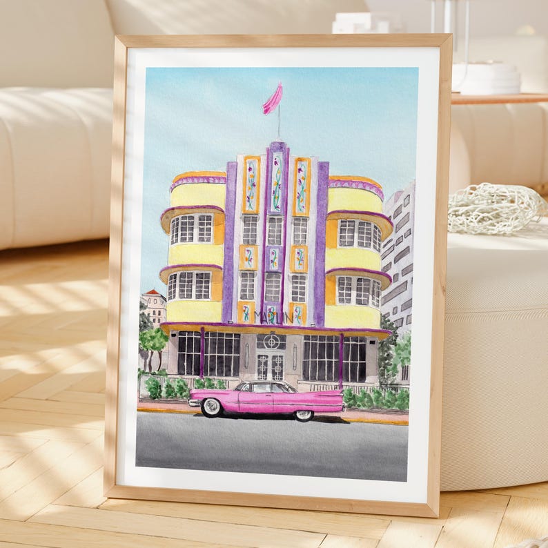 May include: A framed watercolor print depicting the Marlin Hotel in Miami with its yellow and purple art deco facade, featuring a pink vintage car parked in front. The building has multiple windows, a central tower, and a pink flag on top.