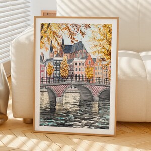 May include: A framed watercolour painting of a European city scene, featuring a bridge over a canal. The artwork shows buildings with red roofs, a church, and autumn leaves in yellow and orange. The bridge is constructed from red brick.