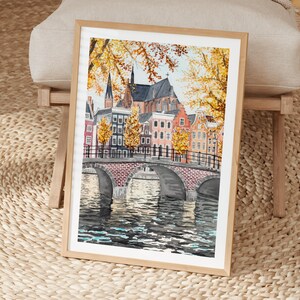 May include: A framed watercolour painting of a European city scene. The artwork depicts a brick bridge over a canal, with colourful buildings and autumn foliage. The painting is in a light wood frame.