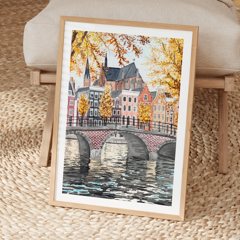 May include: A framed watercolour painting of a European city scene. The artwork depicts a brick bridge over a canal, with colourful buildings and autumn foliage. The painting is in a light wood frame.