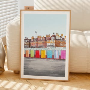 May include: A framed watercolor print depicting a row of colorful beach huts in front of multi-story brick buildings under a pale blue sky. The huts are painted in vertical stripes of orange, green, red, yellow, blue, and pink.