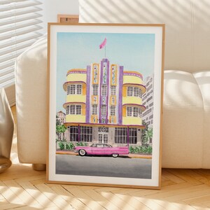 May include: A watercolor art print featuring a yellow and purple Art Deco building with the word MARLIN visible on the facade. A vintage pink car is parked in front of the building on a gray street. The sky is a light blue with a pink flag on top.