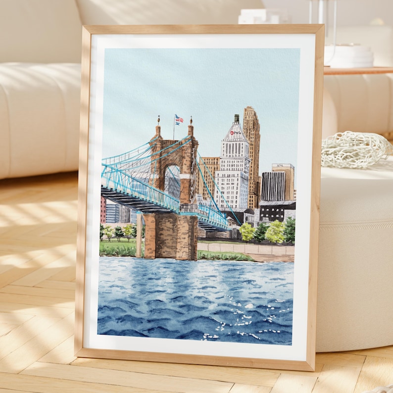 May include: Watercolor painting of a bridge with blue suspension cables and brown stone towers. The bridge spans a blue river, with a cityscape in the background. The artwork is framed in a light wood frame.