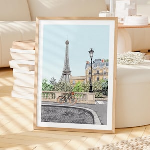 May include: Watercolor print of the Eiffel Tower in Paris, France, with a bicycle parked on a cobblestone street. The artwork features a light blue sky and a classic Parisian street scene, framed in a light wood frame.