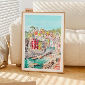 May include: A framed watercolour print depicting a coastal village with colourful buildings in shades of red, yellow, and white, nestled on a rocky cliffside overlooking a turquoise sea with small boats, all set against a clear light blue sky.