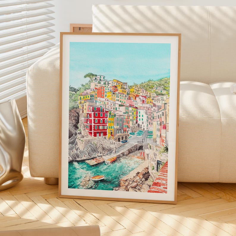 May include: A framed watercolour print depicting a coastal village with colourful buildings in shades of red, yellow, and white, nestled on a rocky cliffside overlooking a turquoise sea with small boats, all set against a clear light blue sky.