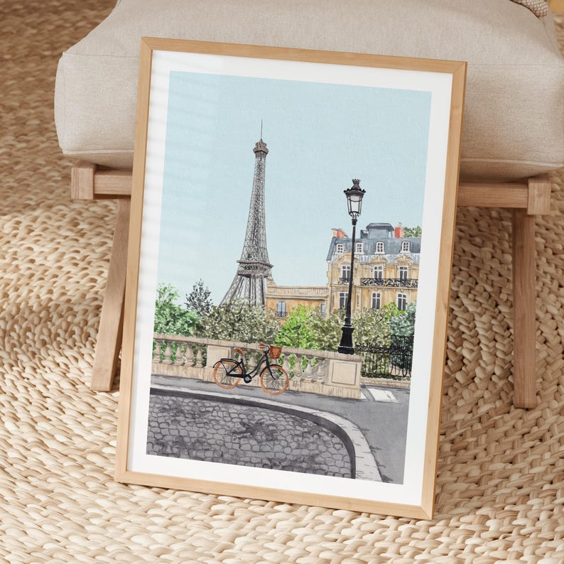 May include: A framed watercolor print of Paris featuring the Eiffel Tower, a bicycle, and a street lamp. The artwork has a light blue sky and a cobblestone street. The frame is made of light-colored wood.