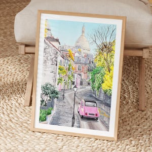 May include: A framed watercolor painting depicting a cobblestone street in Paris with a pink car driving away. The scene features the Sacr&eacute;-C&oelig;ur Basilica in the background, surrounded by stone buildings, green foliage, and yellow autumn trees.