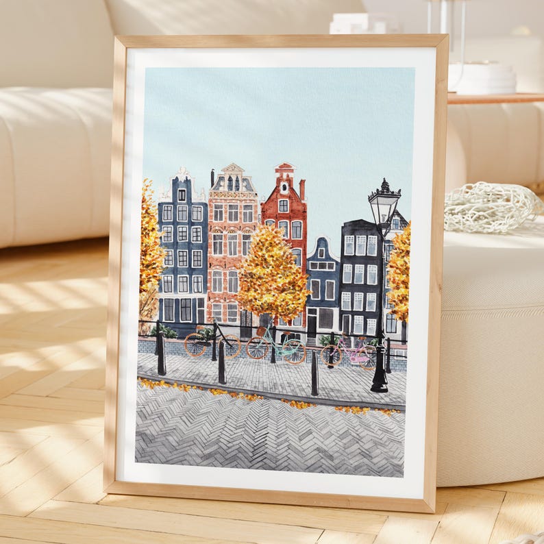 May include: Framed watercolour painting of Amsterdam canal houses. The artwork shows colourful buildings, bicycles, and autumn trees. The sky is light blue, and the cobblestone street reflects the scene.