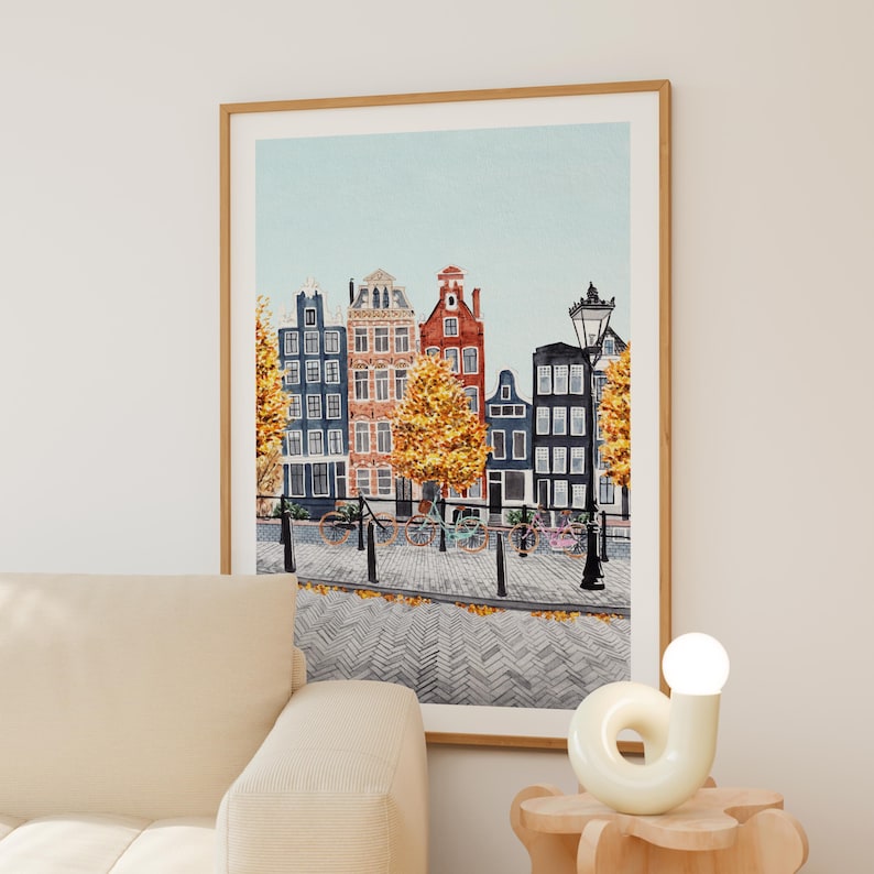 May include: Watercolour print of a European cityscape with colourful buildings, bicycles, and trees. The artwork is framed in a light brown wooden frame. The buildings are painted in shades of blue, orange, and red, with a light blue sky in the background.