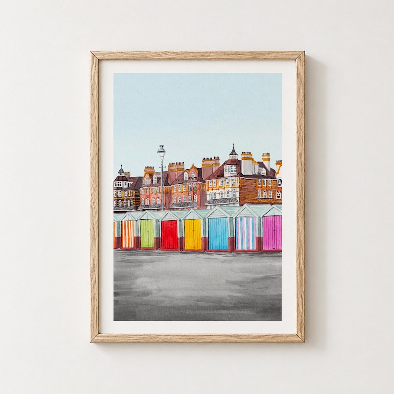 May include: A framed watercolor print depicting a row of colorful beach huts in front of multi-story brick buildings under a pale blue sky. The huts feature vertical stripes in orange, green, red, yellow, blue, and pink, set against a gray ground.
