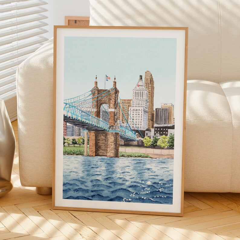 May include: Watercolor painting of a cityscape featuring a blue suspension bridge and buildings. The bridge has blue supports and brown stone towers. The water is blue with light reflections. The artwork is framed in a light wood frame.