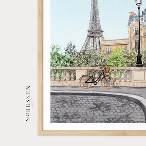 May include: Watercolor print of the Eiffel Tower in Paris, with a bicycle on a cobblestone street. The artwork features a light blue sky and lush green trees, with a decorative stone bridge and buildings in the background. The print is framed in a light wood frame.